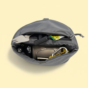 A Campers Pantry CAMPERS MEAL MATE Insulated Meal Pouch in grey nylon. Zippered inner pocket of pouch shown open from above, containing camping utensils including a titanium spork, a Snowpeak Ti Pot, and a Spyderco knife, and Aalto Stove burner. Mesh Pocket on back.